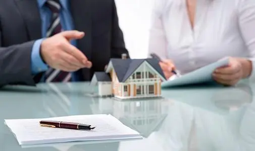 homem de terno e mulher de camisa social sentado em um mesa com uma figura 3d de uma casa e um contrato na mesa.